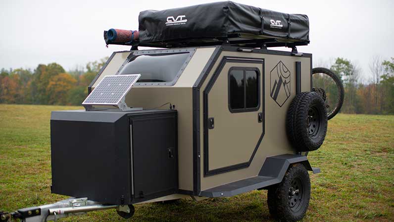 An ALCOM Offroad Oxbow overland camper parked in a field with a solar panel, rooftop tent, and a mountain bike attached to the rear.