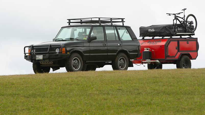 An ALCOM Offroad Spur overland camper being towed by a Range Rover in a large field with a mountain bike on the camper roof.