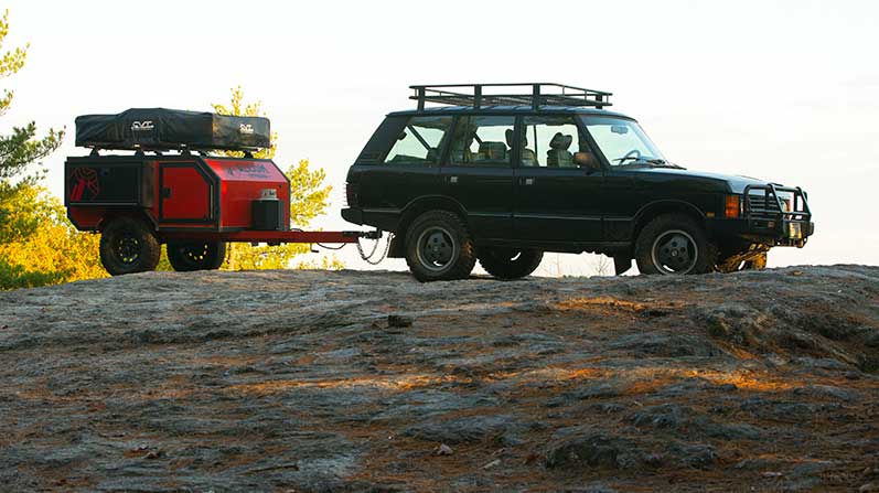 An ALCOM Offroad Spur overland camper towed by a Range Rover on a rocky area.